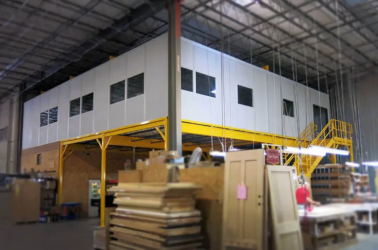 A warehouse interior showcases a modular white office structure elevated on yellow supports. Various materials and wooden items are scattered on the ground floor, while a staircase on the right leads up to the shipping and receiving office area.