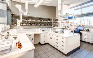 A modern laboratory with pristine white countertops and cabinets features Utah shelving lined with various bottles and containers. Ventilation pipes extend from the ceiling, while a computer setup sits near a window, basking in natural light.
