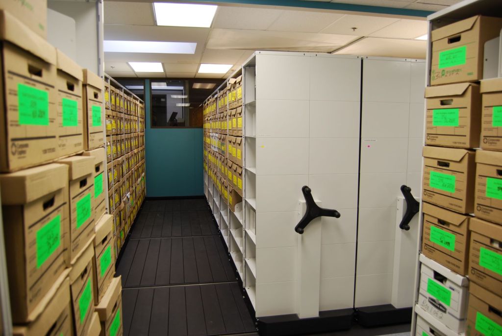 The storage room features shelves of cardboard boxes with green stickers, seamlessly integrating efficient medical records organization. These shelves, part of a movable filing system with hand cranks, offer compact storage under fluorescent lights—a perfect medical records storage solution.
