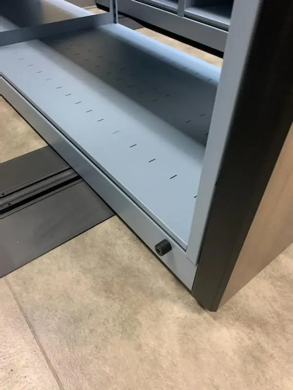 A close-up view of an empty gray metal shelf with perforated holes on a tiled floor hints at its role as mobile shelving for pharmaceutical supplies. A small black knob is visible on the front edge, and a piece of black material lies partially under the shelf.