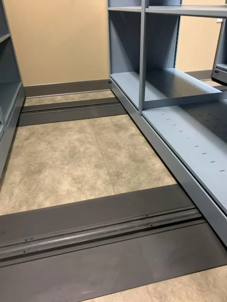 Sliding shelving units on metal tracks installed on a tiled floor, designed for efficient space utilization. Ideal as mobile shelving for pharmaceutical supplies, these shelves are spaced apart to reveal their adjustable positioning and system for compact storage.