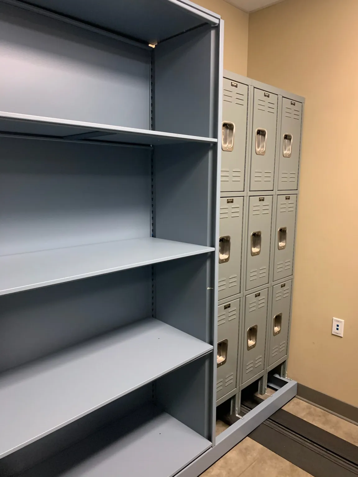 A room with mobile shelving for pharmaceutical supplies and gray lockers. The shelves stand empty, while the lockers are arranged in two columns with ventilation slots. The floor is tiled, and the walls are painted beige.