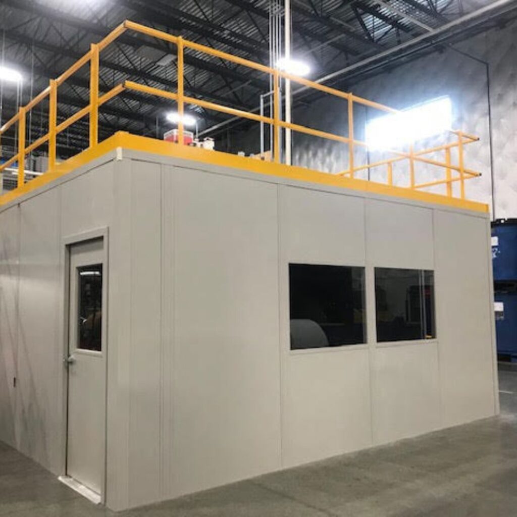 A white modular office with storage roof features a small door and two horizontal windows, all inside a spacious warehouse. The top of the structure boasts a yellow safety railing, perfectly complementing the high ceiling and metal framework of the surrounding space.