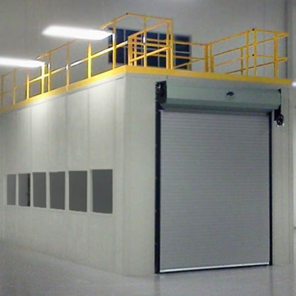 Industrial room with a large closed roller door and a yellow safety railing on top. The area serves as secure storage within a modular building, ideal for specialized operations in warehouse or manufacturing facilities. The walls are white with rectangular windows.