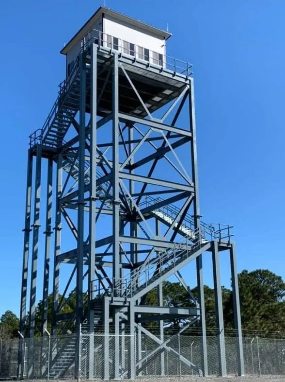 A tall, metal fire watch tower, resembling an operator cab on a 70-foot structure for the US military, features a square observation cabin surrounded by railings. It boasts a spiral staircase and stands against a clear blue sky with a few trees at its base.