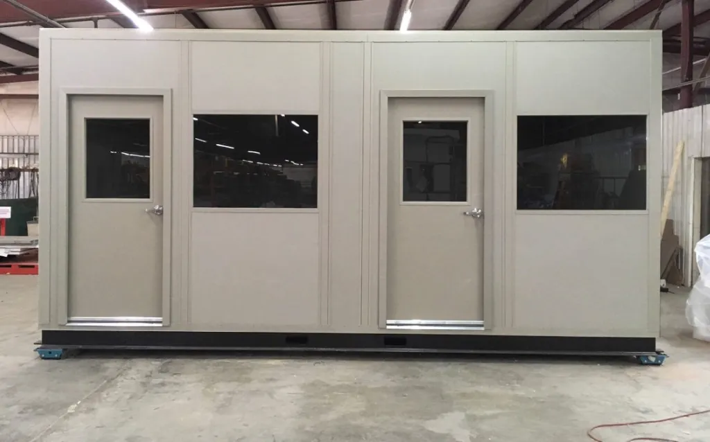 A beige modular booth with two doors and large dark windows stands inside a warehouse, resembling the modern shipping receiving offices. It boasts a flat roof and sits on a platform, with a metal framework visible in the background.