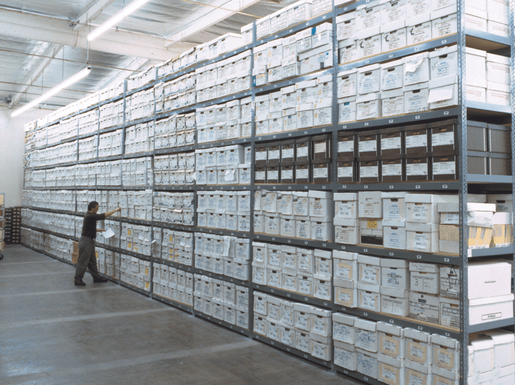 A person examines a vast wall of modular shelving systems filled with stacked white storage boxes in a warehouse. The boxes are labeled, and the shelves stretch from the floor to the ceiling in an expansive space with bright lighting.