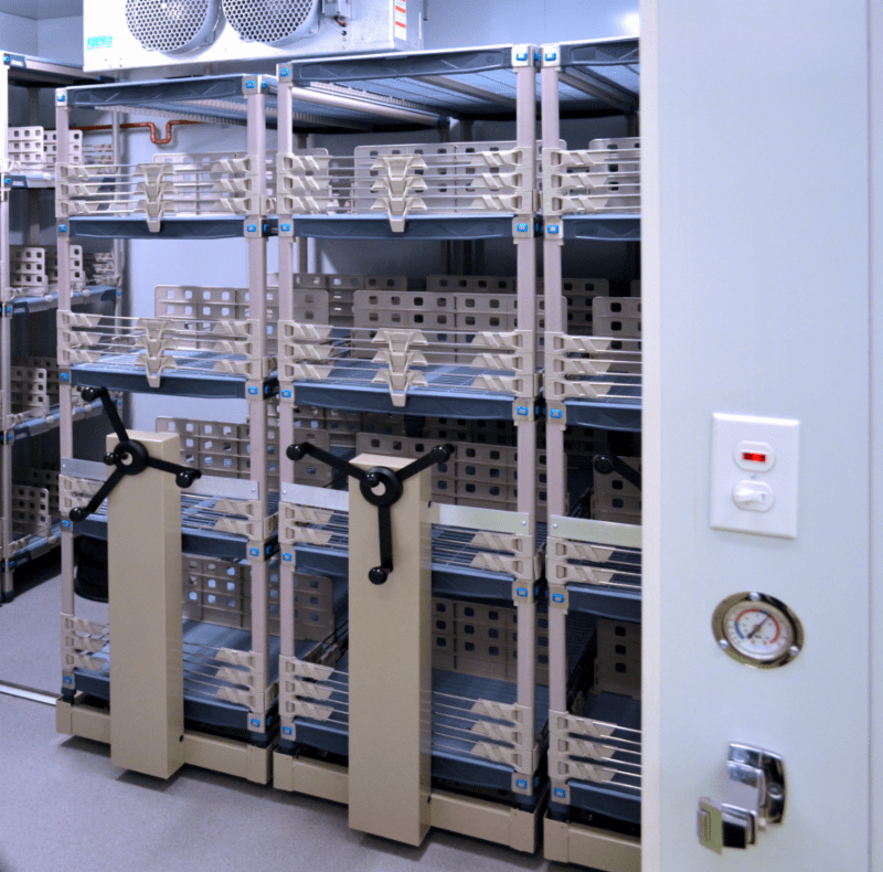 Secure storage room with rows of beige rotary filing cabinets and Mobile High-Density Storage filled with neatly stacked archival boxes. The room features a temperature control unit on the wall, indicating climate-controlled conditions, offering customizable solutions for all your storage needs.