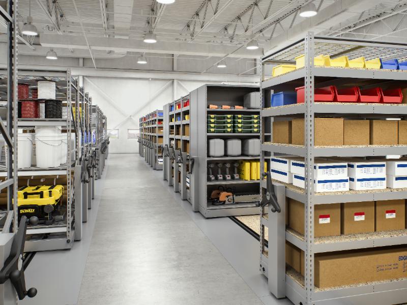 A spacious, secure storage facility with Mobile High-Density Storage solutions features neatly arranged shelves. These shelves hold bins and boxes in vibrant colors like red, yellow, blue, and cardboard brown. Overhead lights illuminate the organized interior to maximize space effectively.