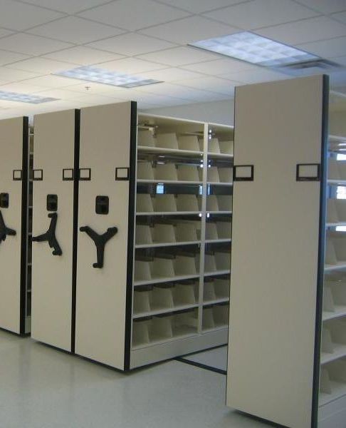 A series of tall, mobile storage shelves in an office setting, reflecting a refined distillery laboratory design. The beige shelves with black handles are easy to move. Most sections remain empty under the glow of fluorescent lighting.