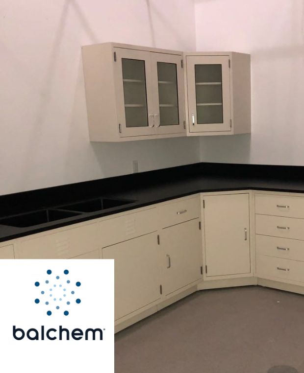 A process research lab corner with off-white cabinets and black countertops. Wall-mounted cabinets have glass doors, showcasing a modern touch. The gray floor complements the sophisticated space, while the Balchem logo, with its blue dotted design, sits proudly in the bottom left corner.