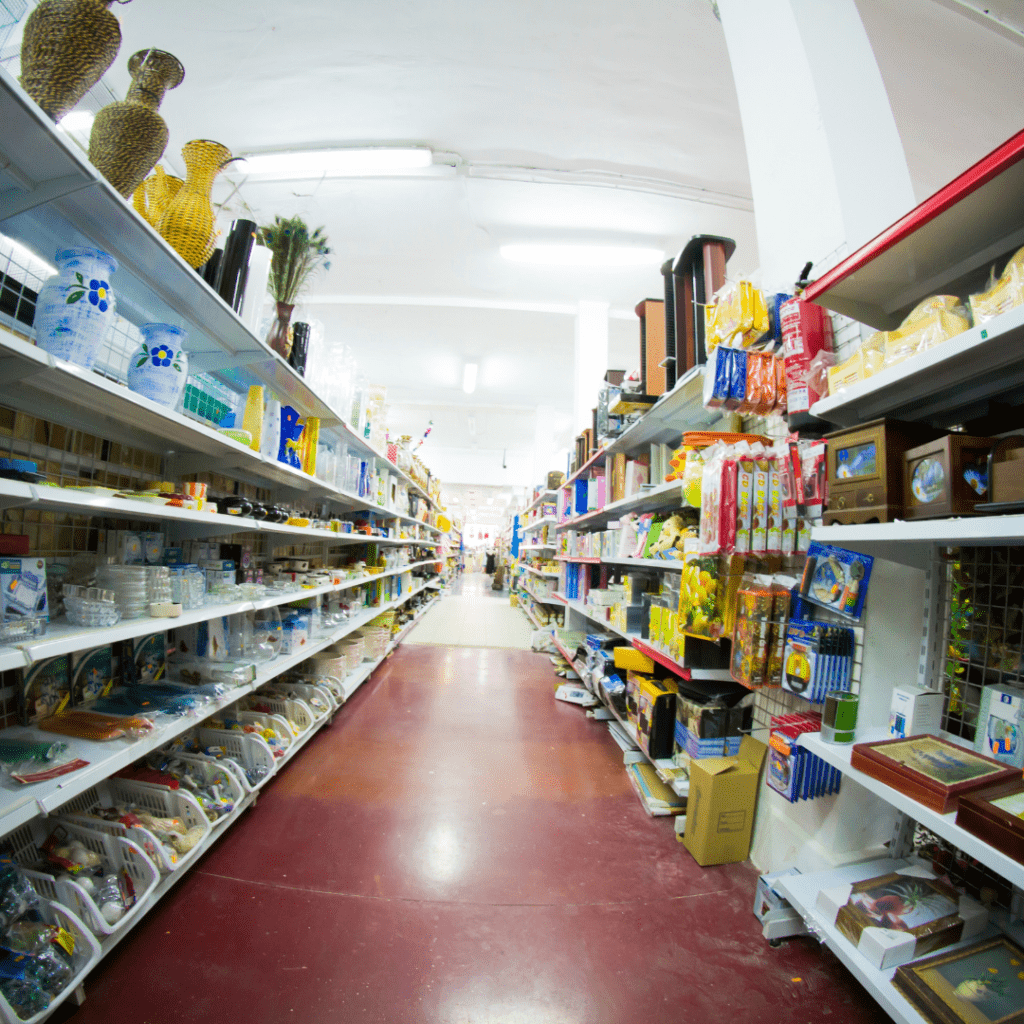 A brightly lit aisle in a store with shelves on both sides brimming with household items like vases, kitchenware, and carpet machines. Cleaning products and packaged goods add to the selection. The store features a red floor and a high white ceiling.