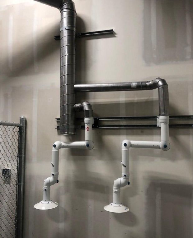 A series of corrosion-resistant metal pipes and ducts mounted on a wall resemble a simplified drawing of a horse. The upper pipes form the head and body, while the lower pipes create the legs. The wall is beige, and theres a chain-link fence on the left.