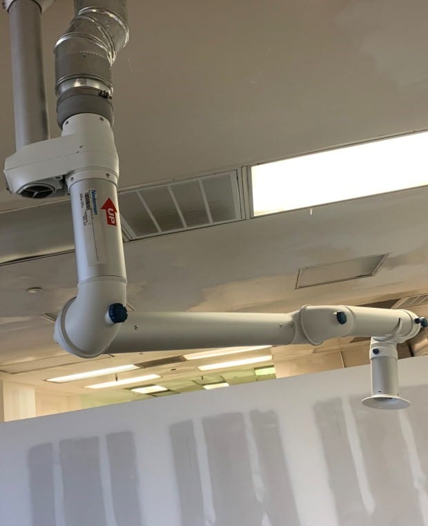 A white adjustable ventilation arm, essential for laboratory fume extraction, is installed on a ceiling, extending towards an open space. The arm features labels and connects seamlessly to ductwork. Bright ceiling lights illuminate the area, highlighting its sophisticated design.