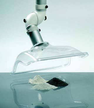 A laboratory fume extraction system with a transparent hood hovers over two piles of powder, one white and one black. The articulated arm adjusts the airflow while positioning the hood above the powders for precise collection, ensuring efficient and safe handling.