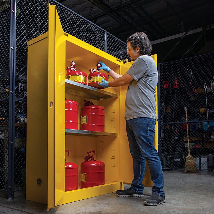 A person in jeans and a gray T-shirt is carefully placing red safety cans into a tall, yellow Justrite safe chemical storage cabinet. The cabinet, nestled within an industrial setting with chain-link fencing in the background, features three sturdy shelves.