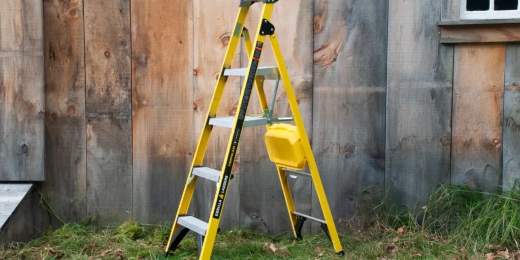 A yellow step ladder with silver steps stands on grass, framed by a weathered wooden wall. To the right, a small window peeks out. Attached to the ladder is a yellow storage tray, embodying the practicality perfect for those seeking functional and stylish ladder images.