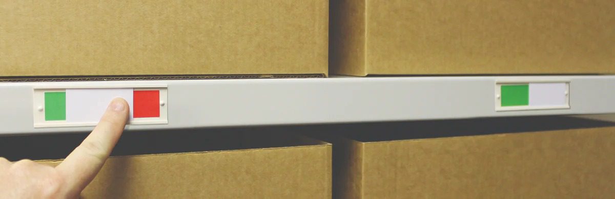 A finger is pressing a red button on a panel mounted on a shelf, possibly triggering the low stock inventory alert. Next to the red button is a green button. The panel, bustling with sticker labels, sits amid brown cardboard boxes and another green button by different boxes.
