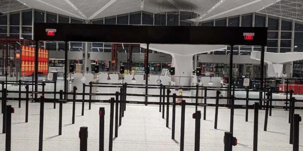 An empty airport check-in area with sleek queue lanes and stanchions offers a sense of order. Large glass windows frame the modern ceiling design, creating a bright space where signs with red exit lights guide travelers, much like flexible storage solutions streamline retail stores.