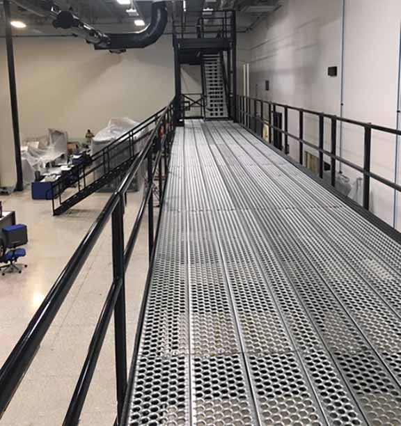 Industrial metal catwalk with black railings, elevated above a bustling workspace. The catwalk extends into the background, lined by a railing. Below, office furniture and covered equipment are visible in the expansive industrial room.
