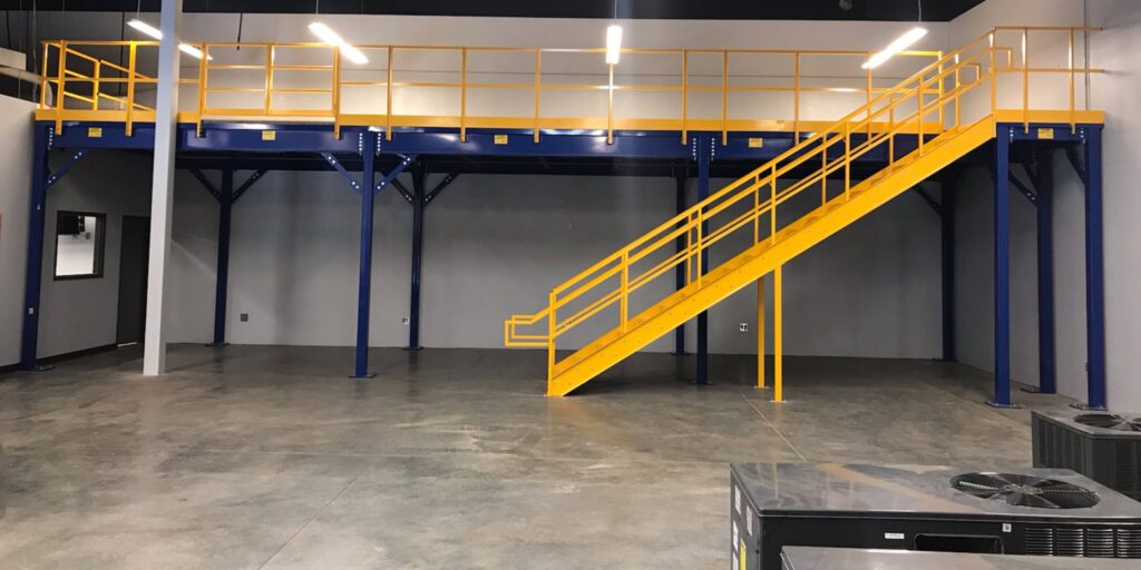 Industrial warehouse interior featuring a striking yellow staircase leading to a blue mezzanine level, the perfect focal point for captivating mezzanines images. The space boasts gray walls, polished concrete floors, and overhead lighting with visible ventilation units in the foreground.