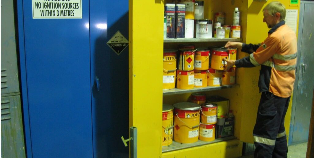 A person in a safety uniform is organizing various paint cans on shelves inside a yellow Justrite Safe Chemical Storage cabinet. Nearby, a blue cabinet features a sign reading No Ignition Sources within 3 Meters.