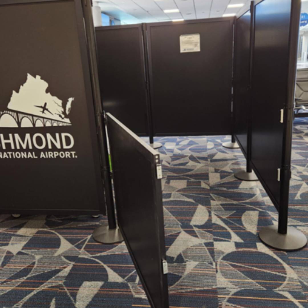A section of an airport terminal features black privacy partitions forming a corridor. One door is slightly ajar, revealing patterned carpeted flooring. Text on a partition reads Richmond International Airport. For those traveling with their pets, this offers a quiet space amid the hustle and bustle.