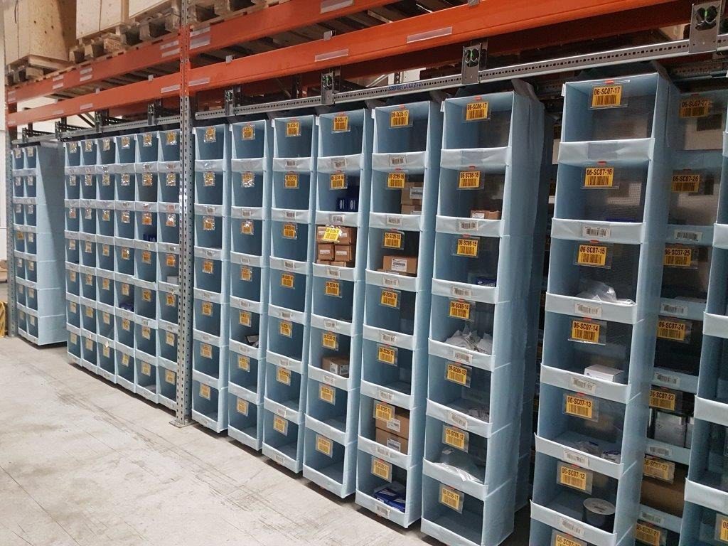 Shelves lined with blue plastic bins reflect a focus on warehouse optimization, as each clearly labeled bin holds small items or boxes. The meticulously organized rows sit against a gray concrete floor, maximizing storage efficiency in the spacious area.