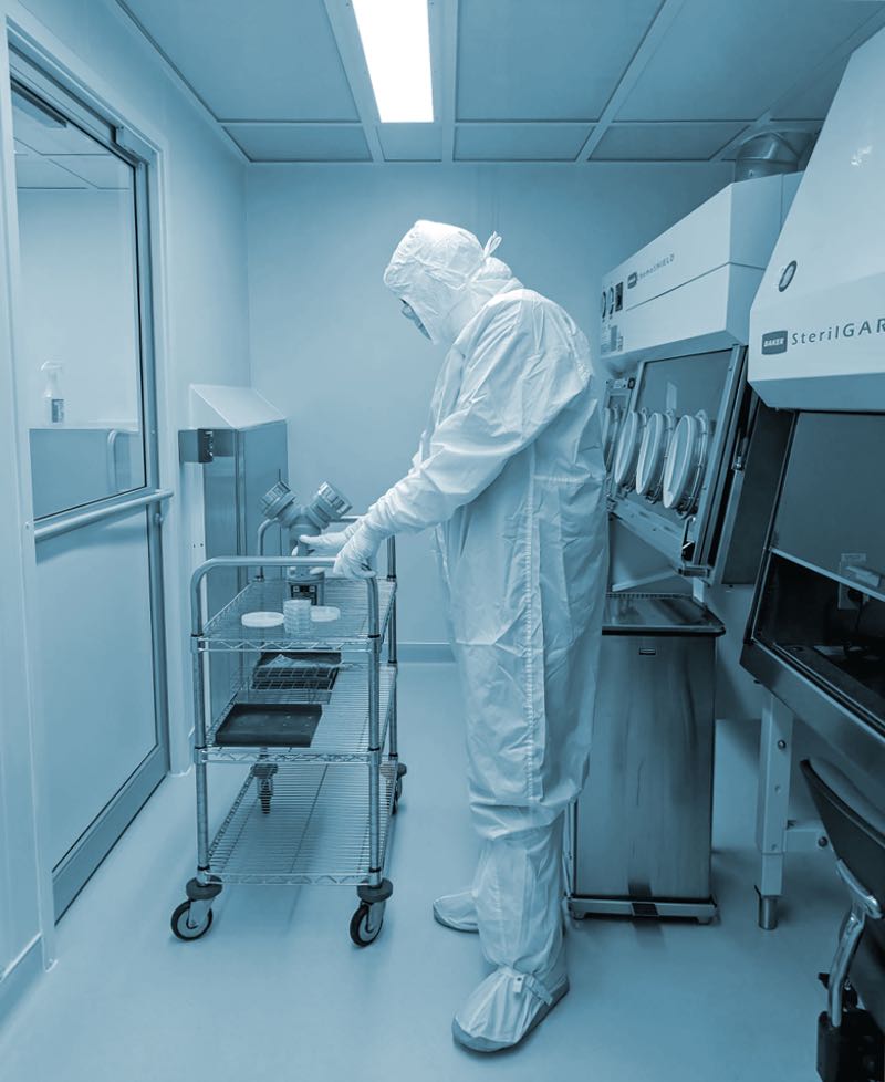 In a sterile laboratory environment, a person in a full-body protective suit and mask meticulously handles items on a metal cart. Surrounded by lab equipment and cleanroom setup, they exemplify the precision and care typical of gowning room essentials.