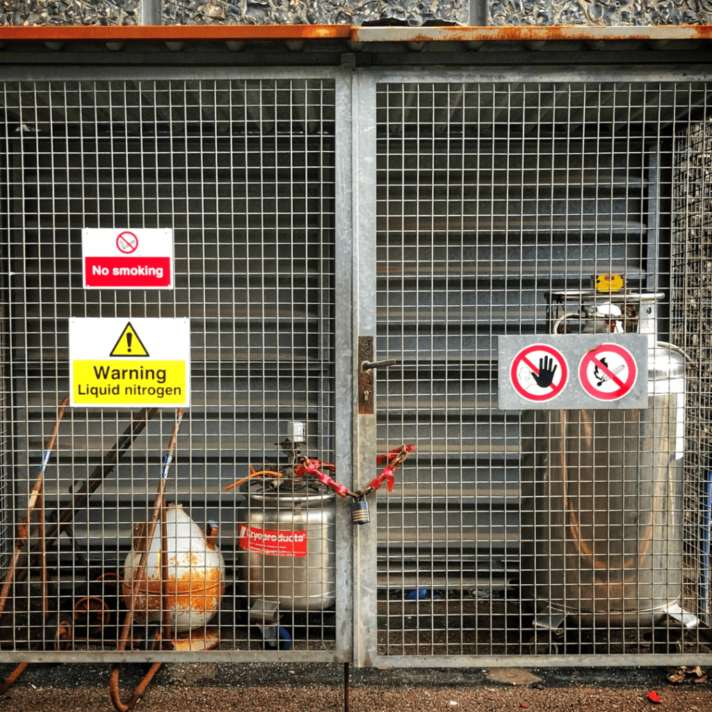 A secured metal cage, monitored by the government department, houses two large metal tanks, one labeled Liquid Nitrogen. Signs on the cage warn against smoking and unauthorized access, emphasizing caution due to the presence of hazardous materials.