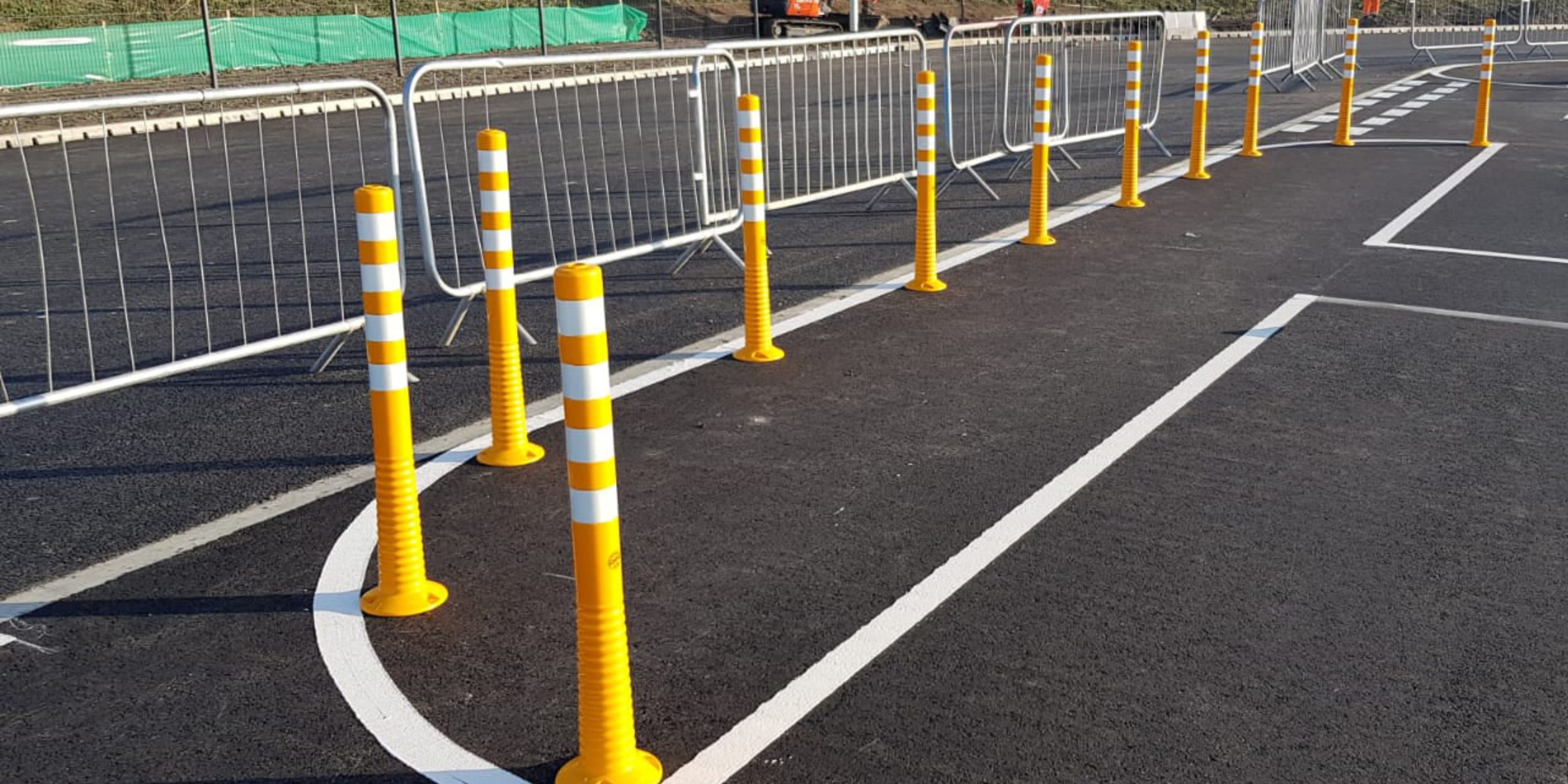Yellow and white striped bollards are strategically lined up along the edge of a newly paved road near metal barriers, enhancing workplace safety. The road markings look fresh, while a green tarpaulin discreetly covers an area in the background.