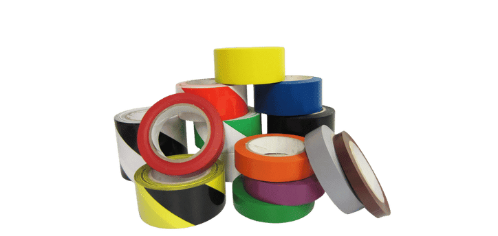 A pile of colorful adhesive tape rolls, vital tools for warehouse organization and safety, are arranged in a semi-circle. The collection includes solid colors like yellow, red, blue, and purple, alongside tapes with safety patterns like black and yellow stripes. The rolls rest at various angles.