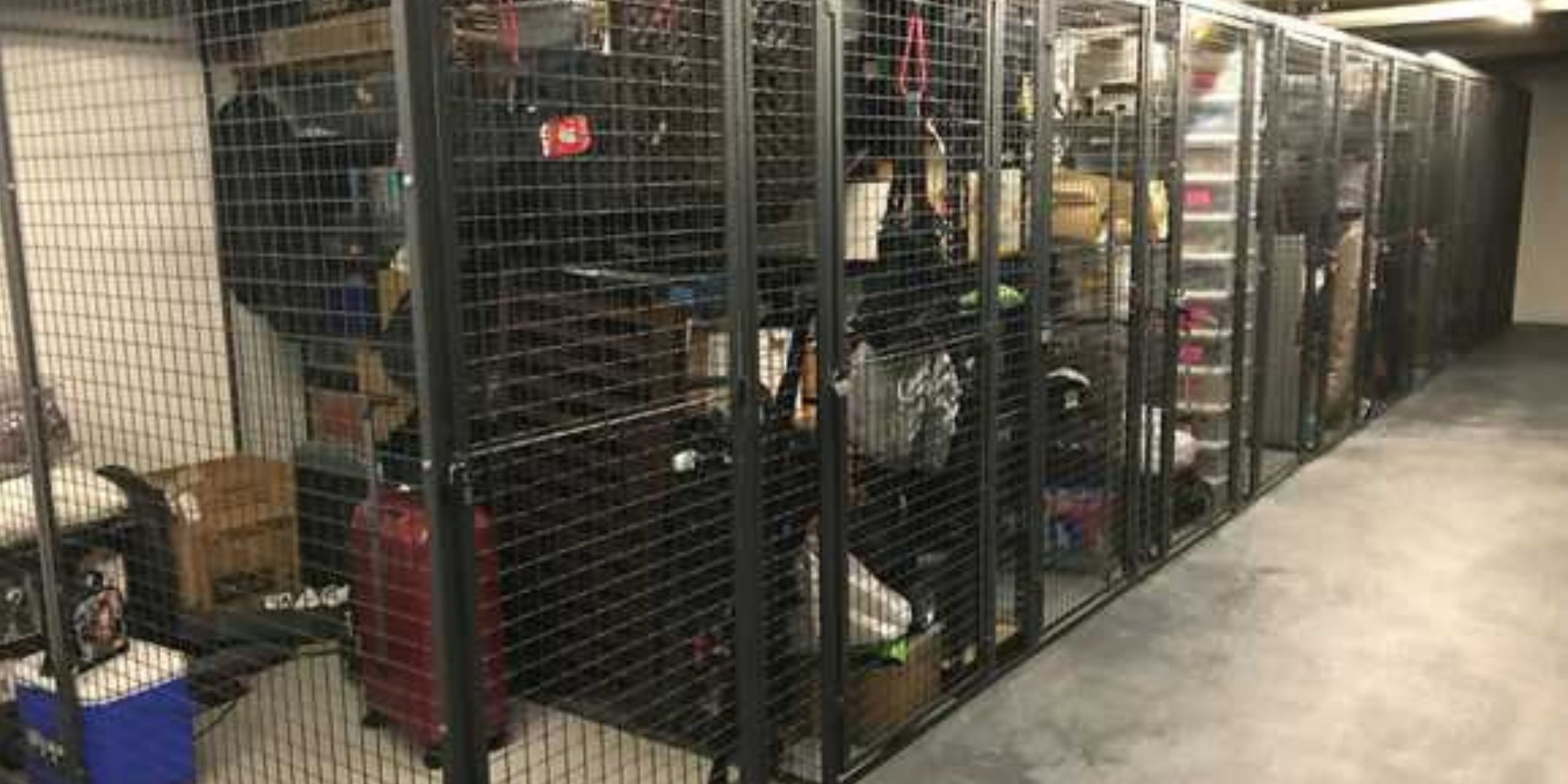 A row of metal storage cages, your ultimate source for tenant lockers, holds boxes, luggage, and plastic containers. These wire tenant lockers are situated in a well-lit, spacious facility with a concrete floor.