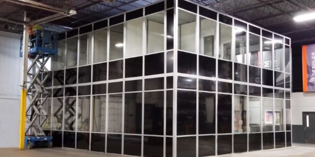 A large, enclosed glass office structure is situated inside a warehouse, featuring black-tinted glass panels reminiscent of Panel Built Images. Reached by a lift, it boasts a high ceiling with visible beams and industrial equipment in the background.