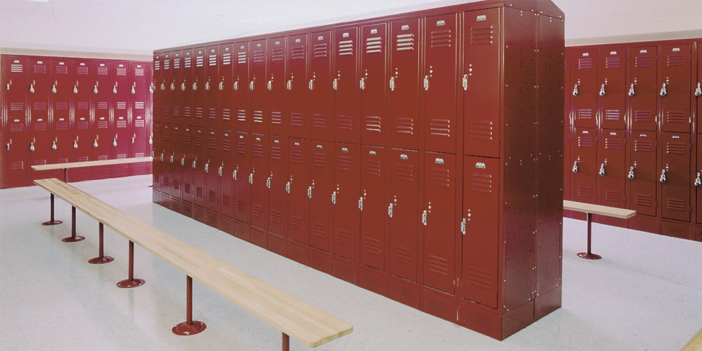 Red metal lockers by Penco Images line the walls of a spacious locker room. Wooden benches sit in front of the lockers, offering seating space. The light-colored floor brightens up the room, creating an inviting atmosphere.