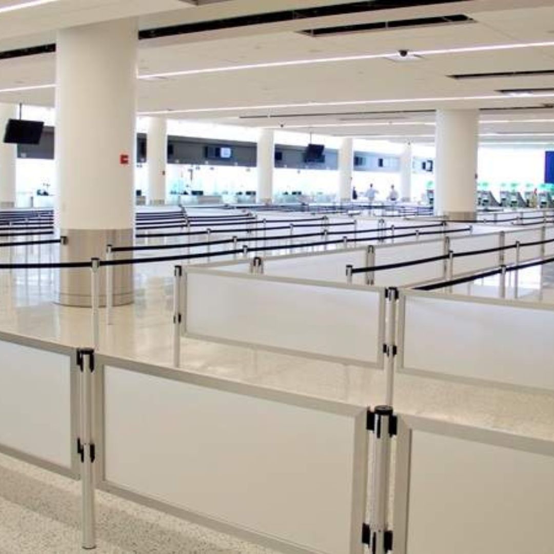 An empty airport security checkpoint area, resembling an office with its white walls and columns, features white barriers and black handrails forming a queue line. The well-lit space includes information screens visible in the background.