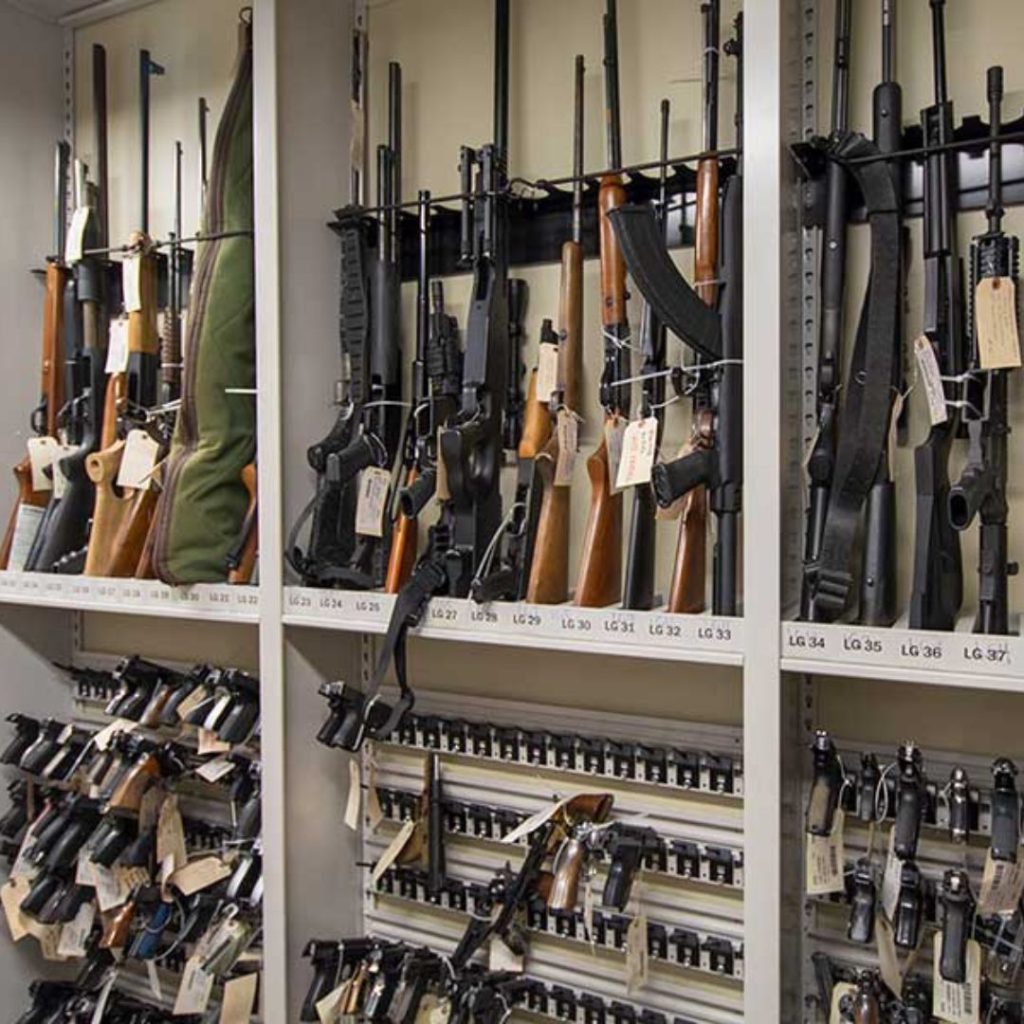 A storage cabinet in the Department: Law Enforcement houses a variety of rifles, shotguns, and handguns neatly organized on shelves. Each firearm bears an identification tag for easy cataloging. Some guns are stacked vertically, while others rest horizontally.