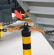 A stairway blocked by a chain barrier attached to a post, marked by a red arrow, highlights efforts to enhance workplace safety. The chain is black and orange while the post is painted yellow with black stripes, giving the setting an industrial or construction-related feel.