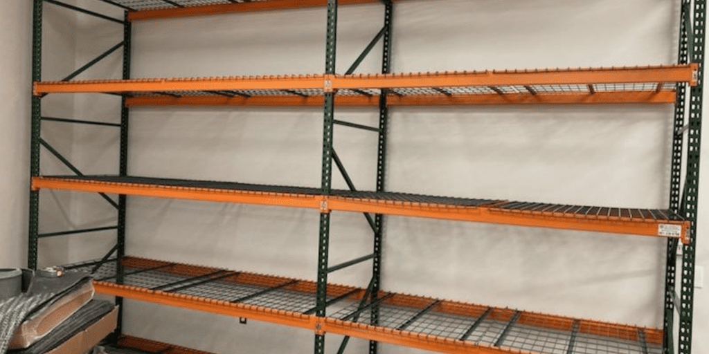 Empty industrial shelving with a metal frame and orange horizontal supports stands against a white wall. The shelves are made of metal grating. Some items are partially visible in the lower left corner.