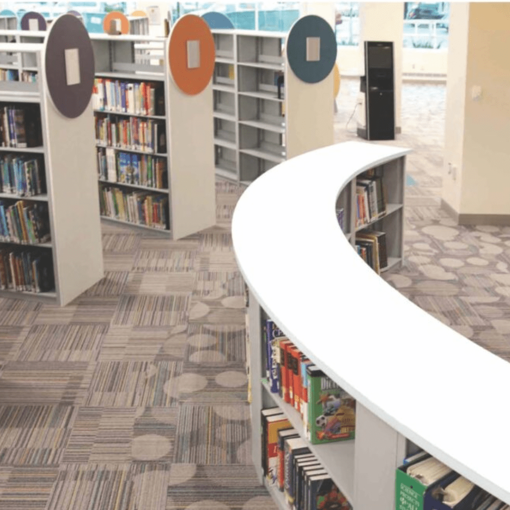 The library interior boasts curved white bookshelves filled with literature, set against patterned carpeting. Tall windows grace the background, while colorful circular signs on shelves guide visitors through this gallery of knowledge.