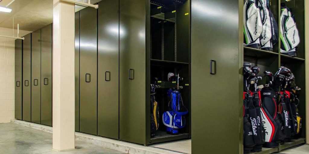 A storage room with large olive-green lockers, reminiscent of Datum storage solutions, is filled with golf bags. Some lockers stand open, revealing a colorful array of blue, black, and red bags. The room features a concrete floor and beige walls.