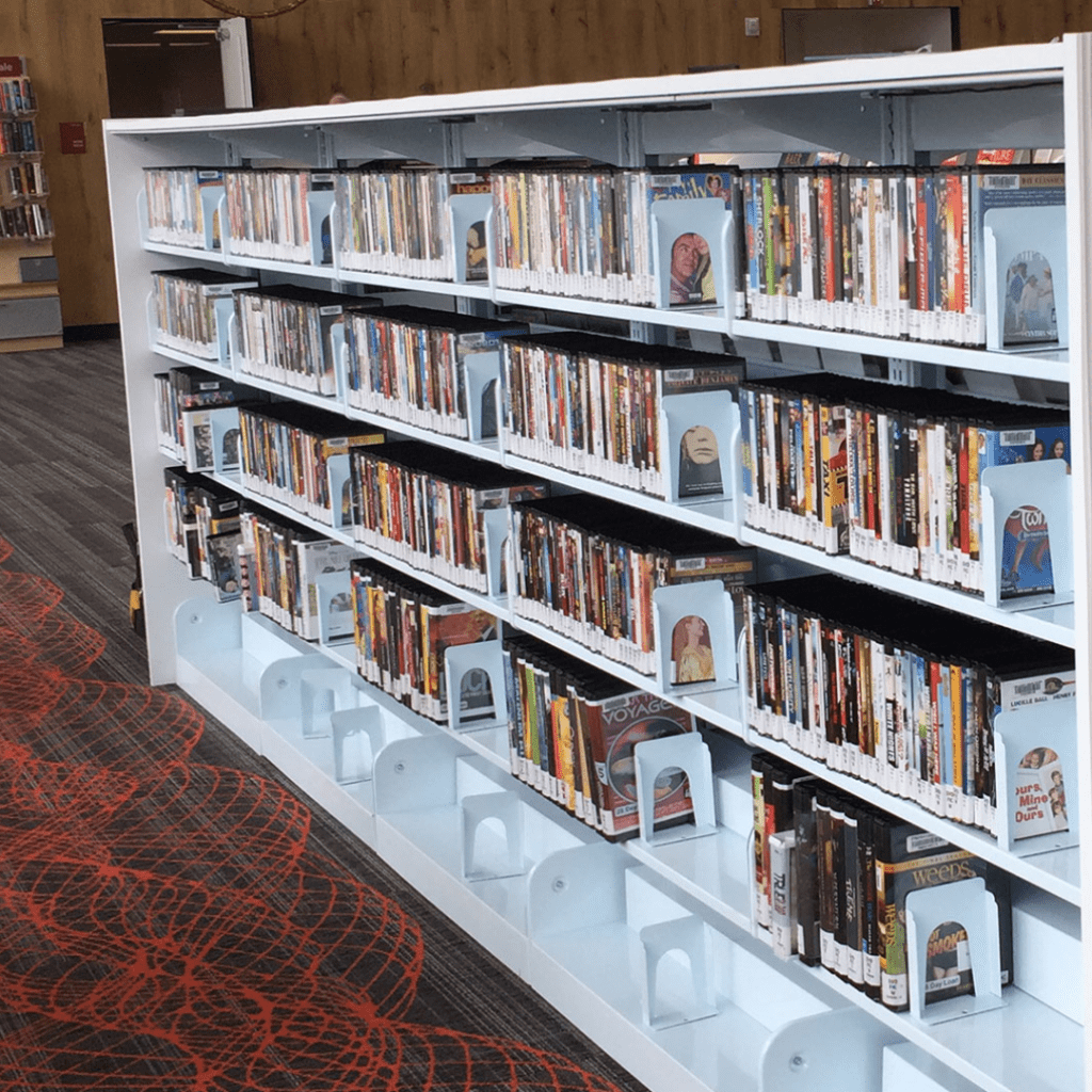 The library shelves, a testament to material handling efficiency, are filled with DVDs in white holders, neatly organized. The shelving unit is white, contrasting elegantly with the dark carpet floor adorned with an orange circular pattern. This corner feels like a literature gallery in its own right.