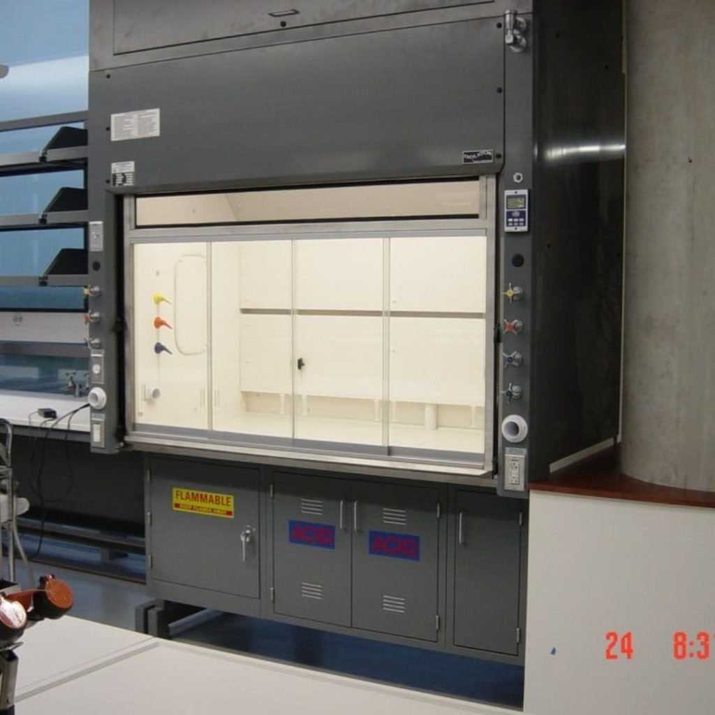 A laboratory fume hood with a transparent sliding sash is pictured, commonly found in the Department of Mining and Gas. It features control knobs on the side and warning labels, including a flammable sign. Shelves and equipment fill the room.