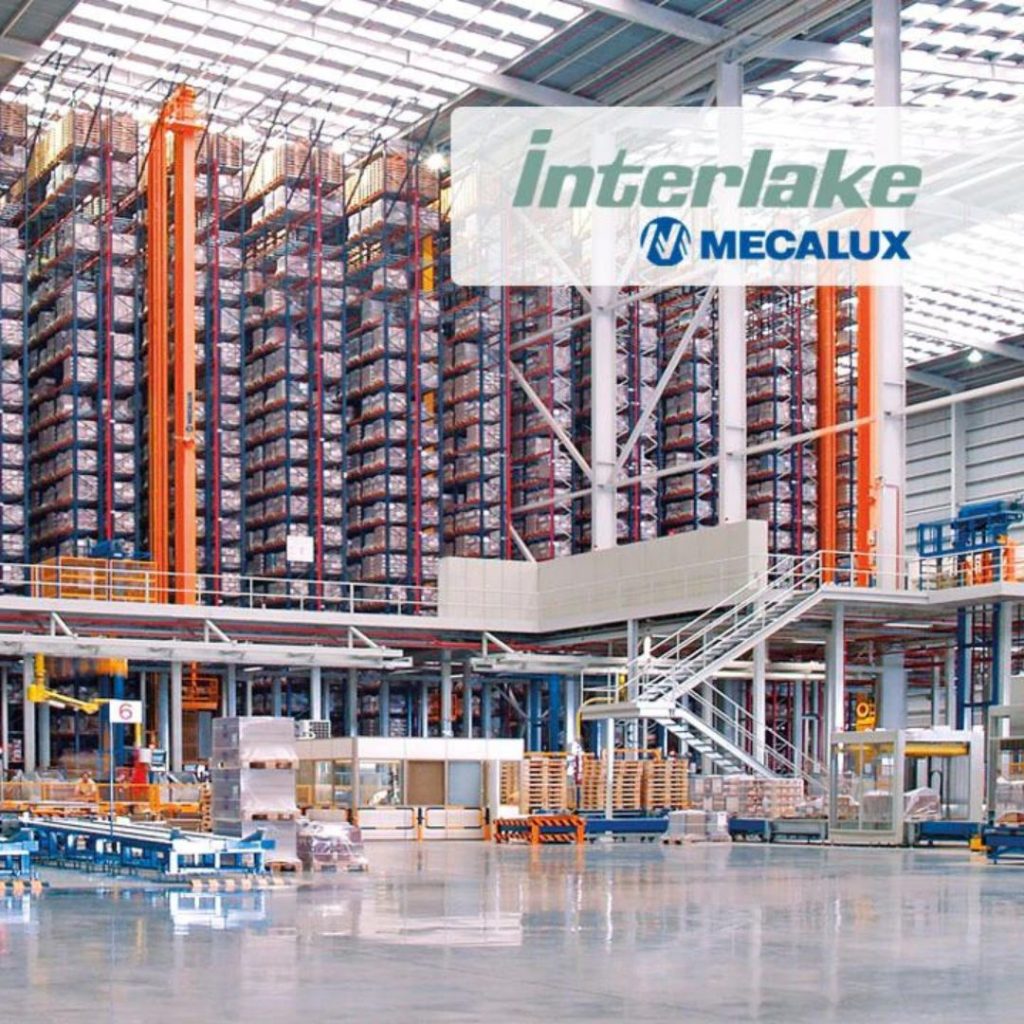 A large warehouse with tall, densely packed storage racks filled with pallets embodies the essence of material handling. Featuring the Interlake Mecalux logo, it boasts bright lighting and industrial equipment like forklifts and conveyor belts, creating a dynamic gallery of efficiency.