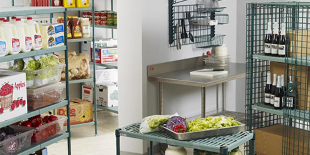 A pantry with metal shelves stocked with milk, canned goods, and fresh produce resembles a scene from Metro Images. A small table holds chopped vegetables, while wine bottles rest on a rack. Cardboard boxes and appliances add to the organized chaos in the background.
