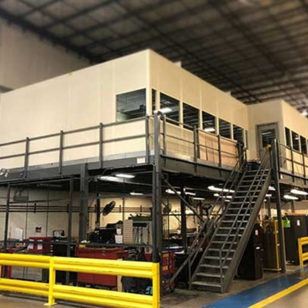 Inside the industrial facility, a large modular structure rises with metal stairs leading to an upper level, characteristic of a Mining and Gas department. Below, equipment and tools rest amid yellow safety rails on the concrete floor under a high ceiling.