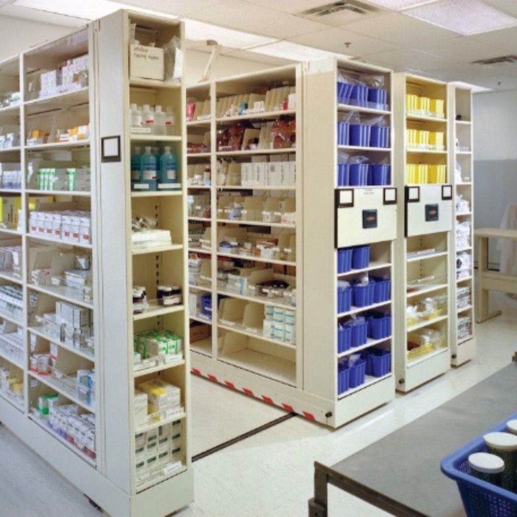 A pharmacy storage area features tall shelves brimming with medications, bottles, and boxes. The well-organized setup resembles a material handling masterpiece, with items neatly placed in clear containers and a blue cart in the foreground. The tidy space could indeed be a gallery of efficiency.