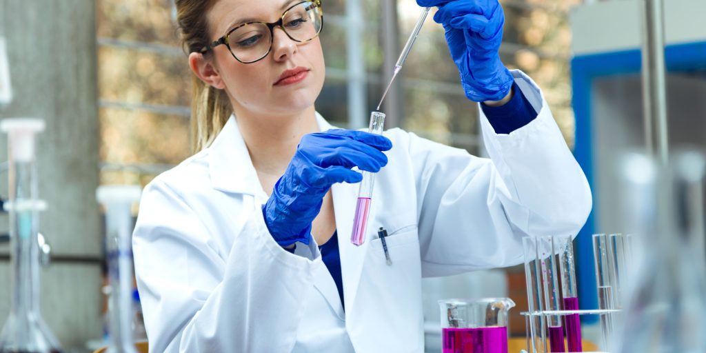 A scientist in a white lab coat and blue gloves carefully pours a pink liquid from a pipette into a test tube. Several glass containers with colorful liquids are on the table, while ISO9001 certified lab instruments fill the background with precision and reliability.