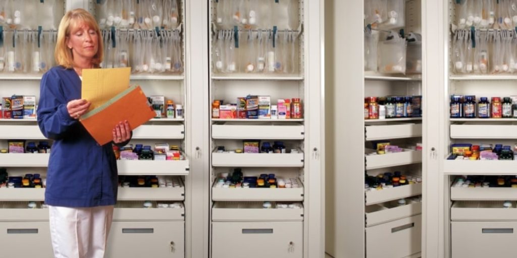 A person in a blue shirt and white pants holds vibrant yellow and orange files, standing twice as confident before tall shelves brimming with medicine bottles and supplies, resembling a scene straight out of one of those classic Times Two Images.