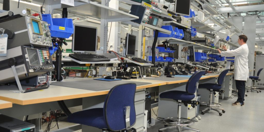 An electronics lab with a row of desks equipped with computers, monitors, and electronic testing equipment. Various tools and blue storage bins are visible on shelves. A person in a white lab coat is standing at one of the workstations.